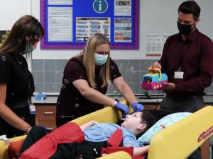 A student is lying on a bed while one adult helps him to access a switch with his head, another adult holds a bubble machine controlled by the switch and a third adult is watching. 