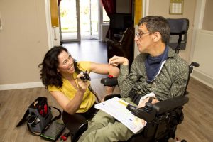 Woman kneeling down smiling and working on the mounting system on a man's wheelchair. The man in the wheelchair is watching and her work and smiling back. 