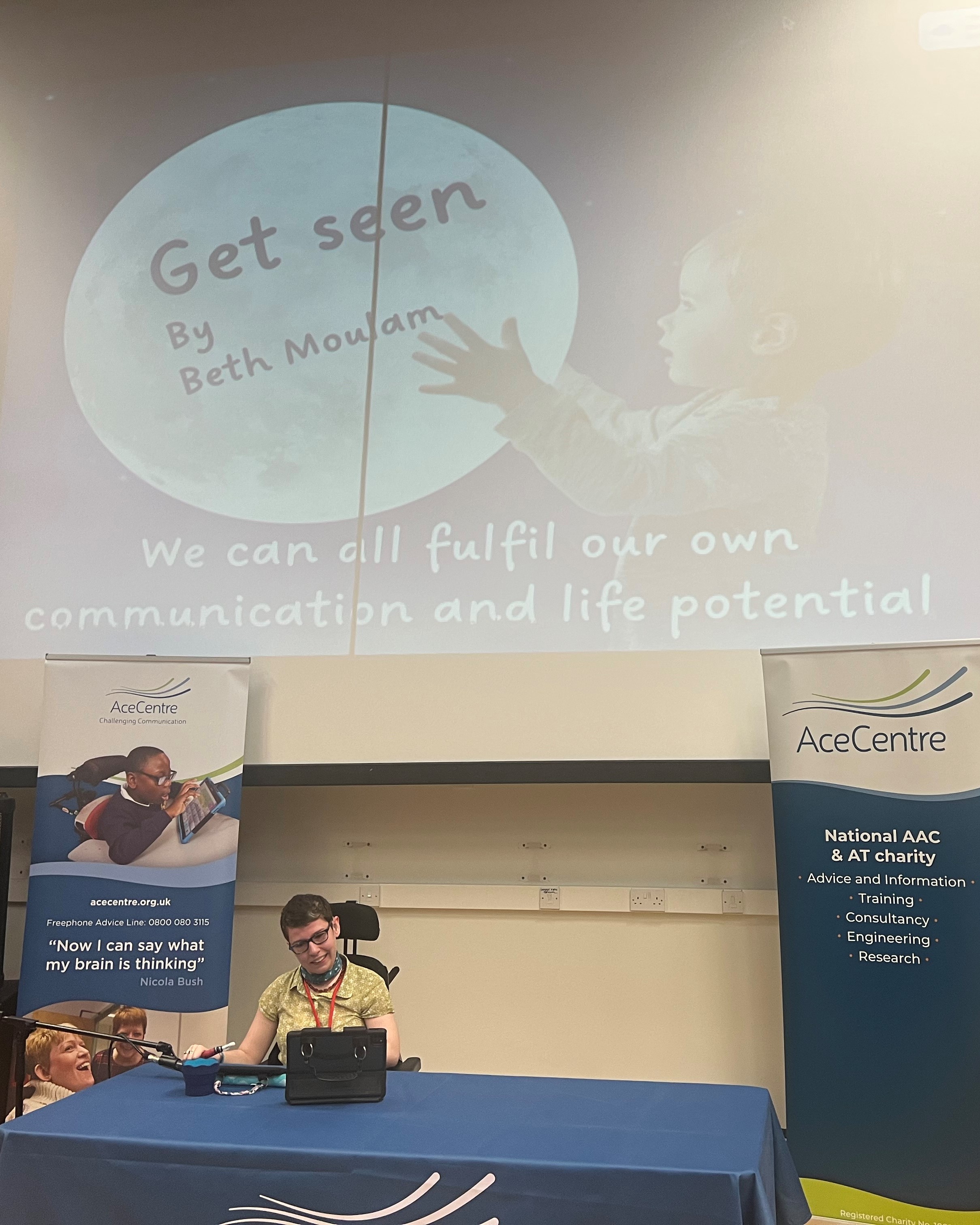 Beth Moulam sitting behind a table presenting her opening speech. There is a power point on display behind her with a picture of a child holding the moon. It says "Get Seen by Beth Moulam We can all fulfill our communication and life potential" 