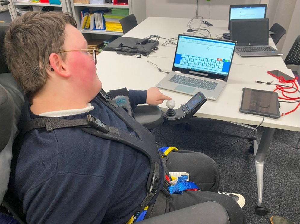 Noah seated in a wheelchair is using an assistive joystick device to interact with his computer. In front of him is a table with a laptop displaying an on-screen keyboard and a tablet with red cables. The setting appears to be an office or classroom with shelves containing colorful folders in the background.